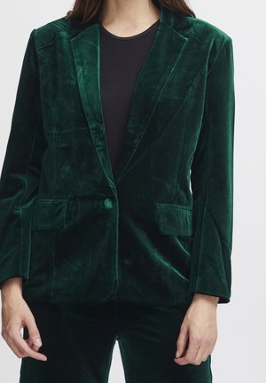 Personne portant un blazer en velours vert foncé avec un seul bouton, des poches à rabat, et une chemise noire en dessous, visible du cou jusqu'aux hanches.