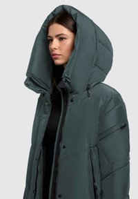 Zelena puffer jakna s velikim, strukturiranim kapuljačom. Sadrži kviltanu teksturu, džepove na zip i dugmad. Mekan, glatki materijal.