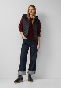 Junge Frau mit langen blonden Haaren, die eine schwarze gepolsterte Weste, einen kastanienbraunen Pullover, geschlossene dunkle Jeans und schwarze Stiefel trägt, und an einer grauen Wand steht.