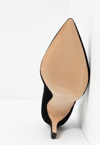Chaussure à talon haut en daim noir à bout pointu, inclinée pour montrer la semelle beige texturée et le talon fin sur fond blanc.