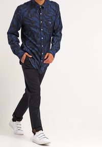 Granatowa koszula z czarnym, wirującym wzorem, długimi rękawami, guzikiem z przodu i kieszenią na piersi. Połączona z ciemnymi spodniami i białymi trampkami.