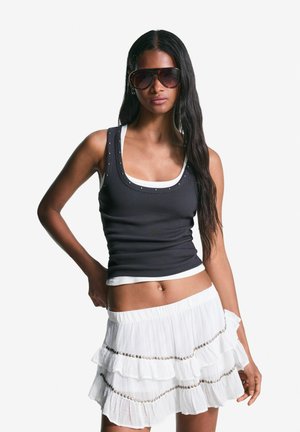 Zwarte geribde tanktop met witte rand en zilveren studs, gecombineerd met een witte rok met ruches en zilveren kralen. Model draagt donkere zonnebril.