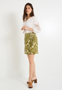 Blusa blanca con mangas bordadas, combinada con una mini falda verde de patrón con diseños de palmeras, y mules marrones de tacón.