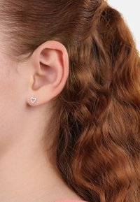 Orecchini a perno a forma di cuore in argento, con un design aperto minimalista, posti su capelli castani ondulati. Gli orecchini sono piccoli e discreti.