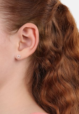 Silberne, herzförmige Ohrstecker mit einem minimalistischen offenen Design, vor gewelltem braunem Haar. Die Ohrringe sind klein und dezent.