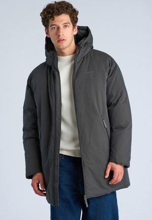 Jonge man met krullend haar die een donkergrijze winterjas met capuchon draagt over een wit overhemd en blauwe jeans, tegen een effen lichte achtergrond.