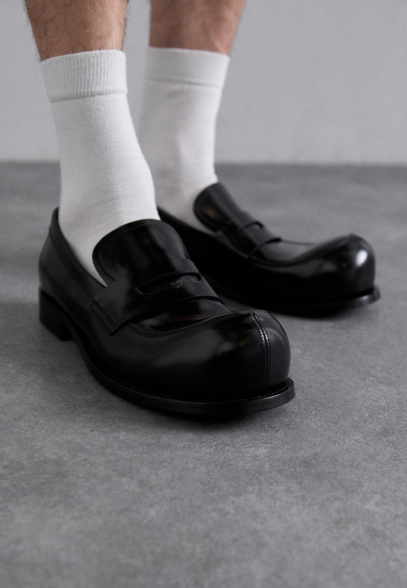 Schwarze Lederloafers mit abgerundetem Zeh, ausgestattet mit einem Vorderen Riemen und einem niedrigen Absatz. Getragen mit weißen Knöchelsocken auf einer grauen Oberfläche.
