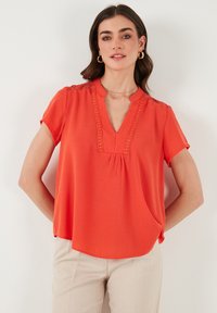 Blusa arancione brillante con maniche corte, scollatura a V, dettagli in pizzo lungo il collo e una vestibilità ampia, abbinata a pantaloni beigi.