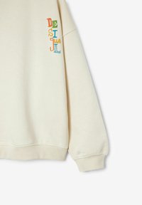 Sudadera de color crema en tejido suave, con el texto bordado "DESIGUAL" en letras coloridas en la manga izquierda y puños acanalados.