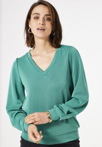 Haut à manches longues en V, de couleur teal uni, fabriqué à partir d'un tissu lisse et léger. Présente des manches froncées et un ourlet ajusté.