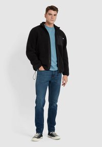 Giacca nera in pile con zip e colletto alto, tasca frontale, indossata sopra una camicia blu e abbinata a jeans blu e scarpe da ginnastica nere.