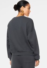 Tummanharmaa huppari, jossa on rento leikkaus, pitkät hihat ja joustavat hihansuut. Varustettu pyöreällä pääntiellä ja pehmeällä, teksturoidulla kankaalla.