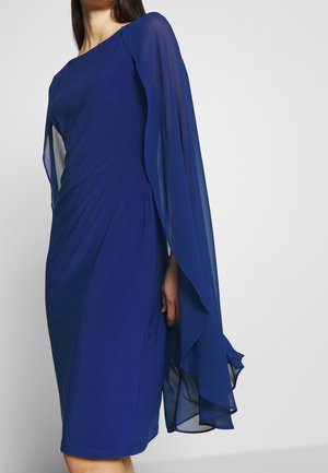 Kobieta ubrana w królewsko niebieską sukienkę do kolan z długimi, przezroczystymi rękawami w formie peleryny i dopasowaną talią, na jasnym, jednolitym tle.