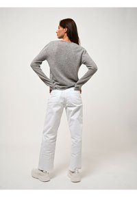 Donna in piedi con le mani sui fianchi, indossa un maglione grigio, jeans bianchi e sneakers bianche, voltata di spalle su uno sfondo semplice.