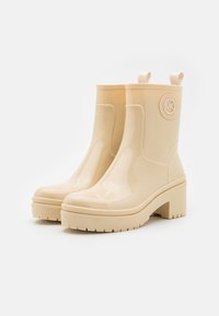 Beige waterdichte enkellaarzen met een glanzende afwerking, chunky hak, een gestructureerde zool en een ronde logo-accent aan de buitenkant.