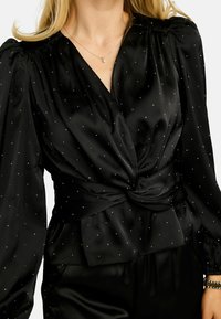 Czarna satynowa bluzka z wiązaniem z przodu, ozdobiona białymi kropkami, z długimi rękawami i marszczonymi ramionami. Gładka faktura i elegancki design.