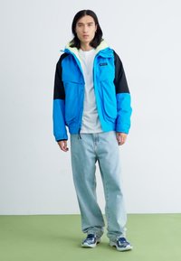 Blauer und schwarzer Mehrfachpanel-Jacke mit Kapuze, weißem Innenfutter; getragen über einem weißen T-Shirt und hellblauen Jeans, kombiniert mit blauen Turnschuhen.