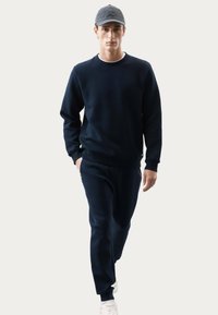 Sudadera navy con textura de espiga y puños acanalados, combinada con joggers navy a juego y zapatillas blancas. El modelo posa de forma casual.