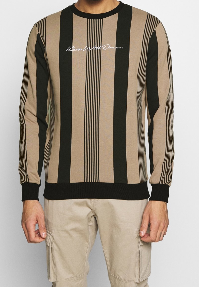 Chemise à manches longues beige et noire à rayures verticales avec le texte "Kings Will Dream", portée avec un pantalon cargo beige.