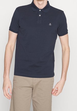 Mann trägt ein marineblaues kurzärmeliges Poloshirt mit zwei Knöpfen und beige Hose, steht vor einem einfarbigen Hintergrund.