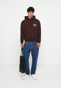 Bruine hoodie met een voorkleurige zak en logo, gecombineerd met blauwe denim jeans en witte sneakers. Zwarte tote bag met logo in hand.