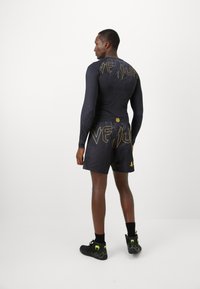 Mann in schwarzer Venum-Sportbekleidung mit goldener Schrift und Schlangendesign, der vor einem schlichten weißen Hintergrund steht, von hinten gesehen.