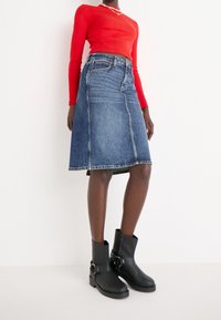 Dżinsowa spódnica w kształcie litery A z kontrastowymi szwami, w jasnoniebieskim kolorze, o średniej długości, zestawiona z dopasowaną, czerwonym długim rękawem bluzką i czarnymi botkami za kostkę.