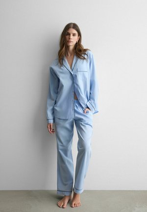 Donna in piedi a piedi nudi contro un muro, indossa un pigiama azzurro chiaro con bottoni e piping nero, una mano in tasca, posa rilassata.