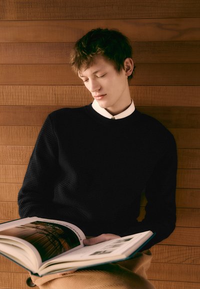 Pull en tricot noir texturé sur une chemise blanche, avec un livre de couleur claire tenu contre un arrière-plan en bois. Pose de lecture, expression détendue.