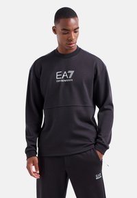Sudadera negra con cuello redondo, logo en el pecho y detalle de panel contrastante. Tejido suave y mangas largas, combinada con pantalones a juego.