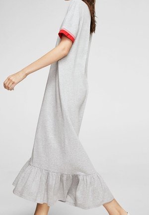 Kobieta ubrana w jasnoszarą sukienkę do kostek z krótkimi rękawami wykończonymi czerwonym paskiem i falbaną u dołu, idąca na tle jednolitego tła.