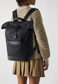 Mochila negra tipo roll-top con correas ajustables, hecha de tejido texturizado y con detalles de cuero, que cuenta con un logo en la parte delantera y hardware resistente.