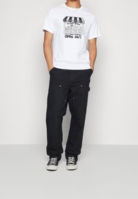 Vit t-shirt med svart grafiskt tryck som visar en växtbutikdesign, ihop parat med svarta löst sittande byxor och svarta sneakers.