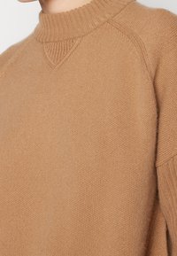 Pull en tricot couleur camel avec un col montant côtelé, des manches raglan, et une finition douce et texturée. Présente un détail discret en point de V.