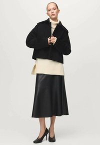 Abrigo negro corto con solapas anchas, superpuesto sobre un suéter de cuello alto de canalé color crema. Combinado con una falda larga de cuero negra y tacones altos.