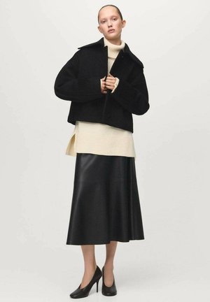 Abrigo negro corto con solapas anchas, superpuesto sobre un suéter de cuello alto de canalé color crema. Combinado con una falda larga de cuero negra y tacones altos.
