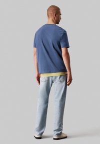 T-shirt blu a maniche corte sovrapposto a una maglietta gialla, abbinato a jeans azzurri chiari. Il modello è visto da dietro, indossando scarpe bianche.