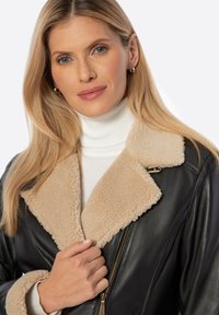 Zwarte leren jas met een beige schapenvacht kraag, ritssluiting, ribbelige witte coltrui eronder, gladde textuur en eenvoudige gouden hardware accenten.