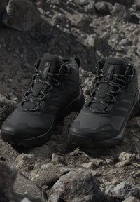 Par sorte Adidas Terrex vandresko med snørebånd på stenfuldt, ujævnt terræn udendørs.