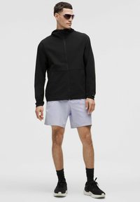 Man in een zwarte zip-up hoodie, lichtgrijze shorts, zwarte sneakers en een zonnebril, staand tegen een effen lichte achtergrond.