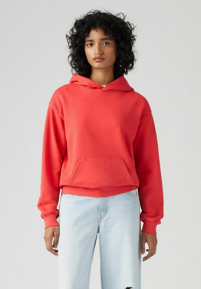 Felpa rossa con cappuccio e tasca frontale, realizzata in materiale morbido. Indossata con jeans azzurri strappati per un look casual.