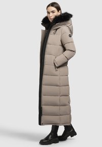 Bezs színű, párnázott hosszú kabát fekete cipzárral, oldalsó zsebekkel és levehető fekete prémes kapucnival. Különleges mintája steppelt textúra, és egyenes sziluettel rendelkezik.