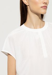 Weiße Bluse mit kurzen Ärmeln und rundem Halsausschnitt, mit einer Vorderlegung und weichem, leichtem Stoff mit glatter Textur.