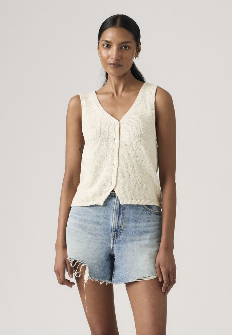 Woman wearing a sleeveless cream buttoned knit top and light blue distressed denim shorts standing against a plain background.