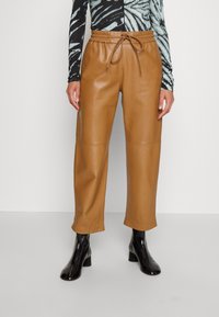 Person iført brune læder cropped bukser med snoretræk i taljen og sorte patent ankelstøvler på en ensfarvet baggrund.