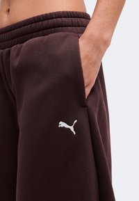 Άτομο φοράει σκούρο καφέ φόρμα Puma με το χέρι στην τσέπη, δείχνοντας το ελαστικό κορδόνι και το λογότυπο Puma στο μηρό.