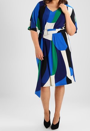Vrouw draagt een knielange jurk met blauwe, groene, zwarte en witte geometrische patronen, een tailleband en zwarte pumps met spitse neus.