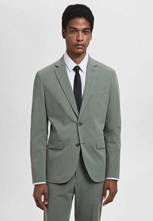 Giacca da suit grigio chiaro con rever a punta e due bottoni. Presenta tasche frontali e un tessuto strutturato, abbinata a una camicia bianca e una cravatta nera.