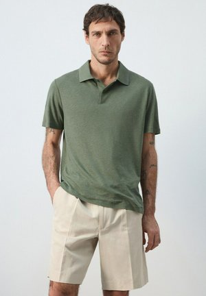 Mann mit kurzen dunklen Haaren, der ein olivgrünes kurzärmeliges Poloshirt und beige knielange Shorts trägt, steht mit den Händen in den Taschen.