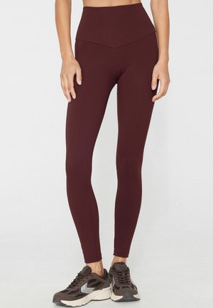 Mujer con leggings de cintura alta de color marrón oscuro y zapatillas de correr marrón oscuro, de pie frente a un fondo liso.